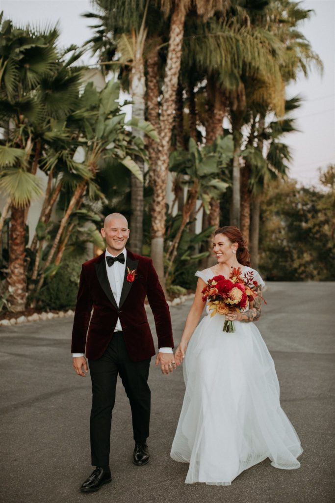 bride in modern asymmetrical dress and bright bridal bouquet stands with groom in red velvet suit jacket in front of mid-century Capri Hotel in Ojai