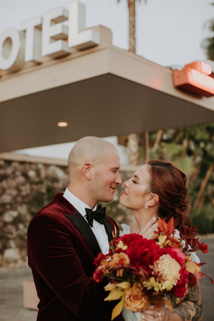 bride in modern asymmetrical dress and bright bridal bouquet stands with groom in red velvet suit jacket in front of mid-century Capri Hotel in Ojai