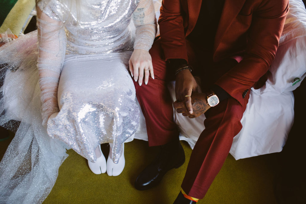 bride in silver wedding dress sits on bed with groom in red suit