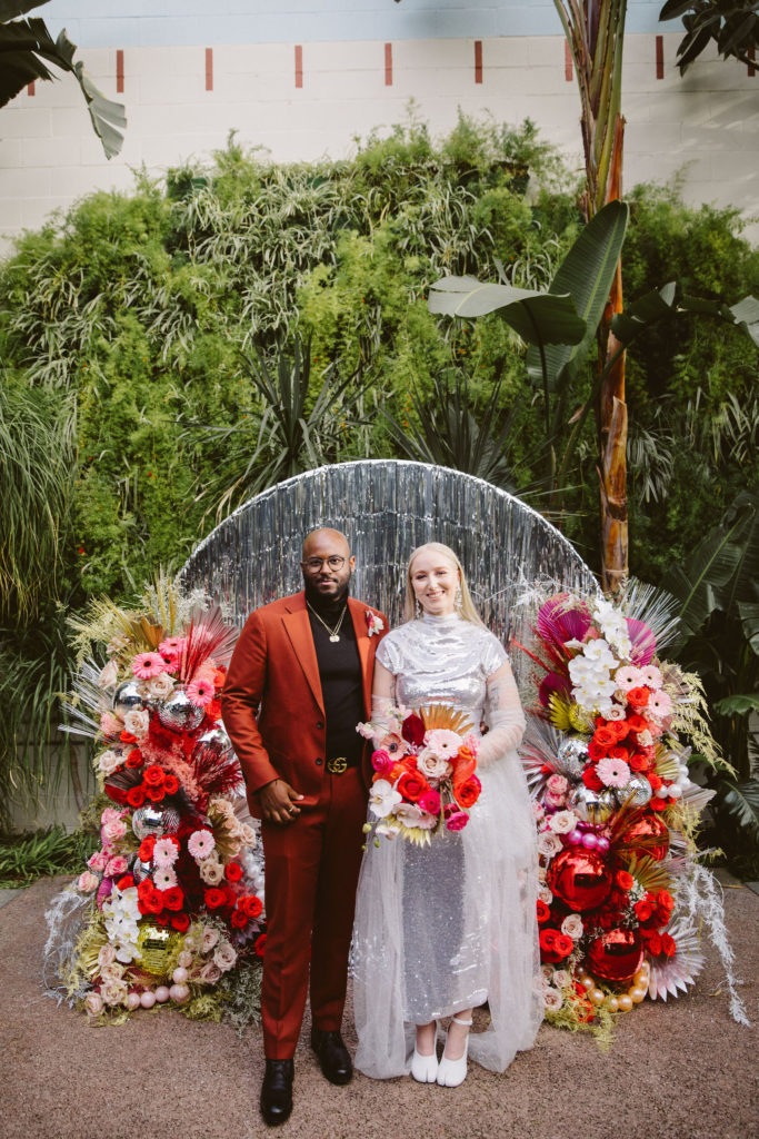 bride in silver wedding dress stands with groom in red suit in front of a silver tinsel ceremony background with red and pink florals