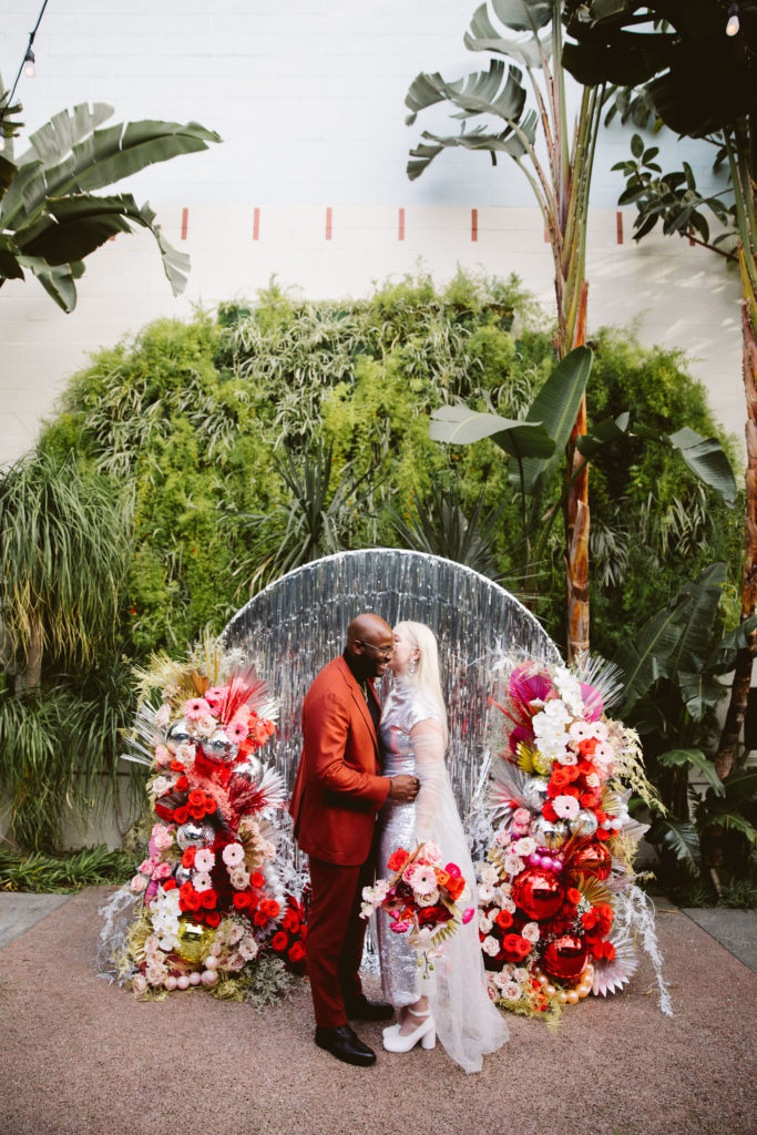 bride in silver wedding dress stands with groom in red suit in front of a silver tinsel ceremony background with red and pink florals