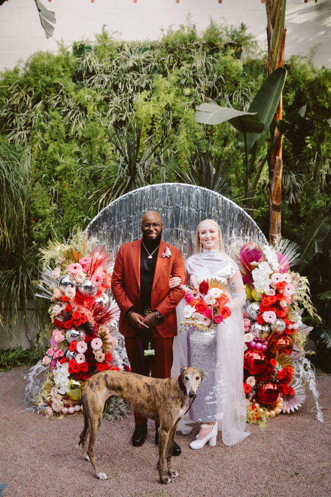 bride in silver wedding dress stands with groom in red suit with their dog in front of a silver tinsel ceremony background with red and pink florals