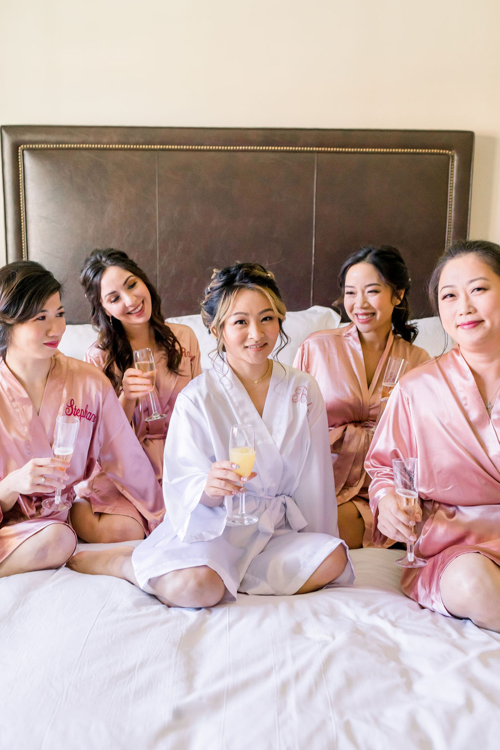 bride in white satin robe sits on bed with bridesmaids in personalized pink satin robes