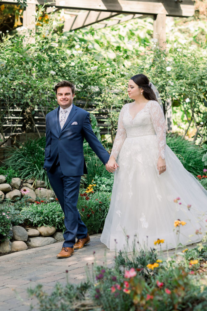 bride in long sleeve lace wedding dress holds hands with groom in blue suit and floral tie