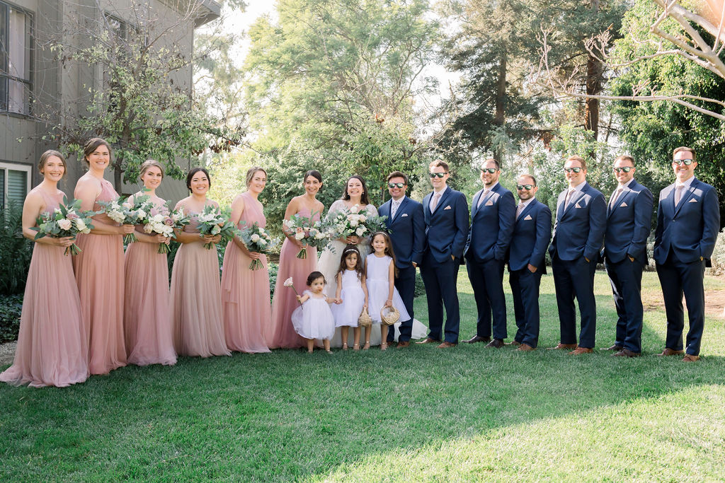 wedding party photo with bridesmaids in light pink dresses and groomsmen in blue suits with floral ties plus three flower children in white dresses