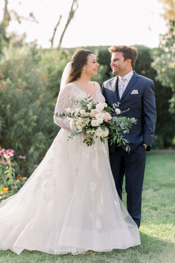 bride in long sleeve wedding dress with floral lace appliqué and sweetheart neckline stands with groom in blue suit and floral tie at Maravilla Gardens