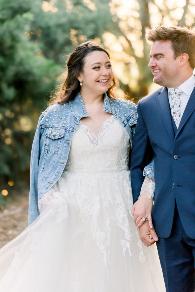 bride in long sleeve wedding dress with pearl embellished denim jacket walks with groom in blue suit and floral tie