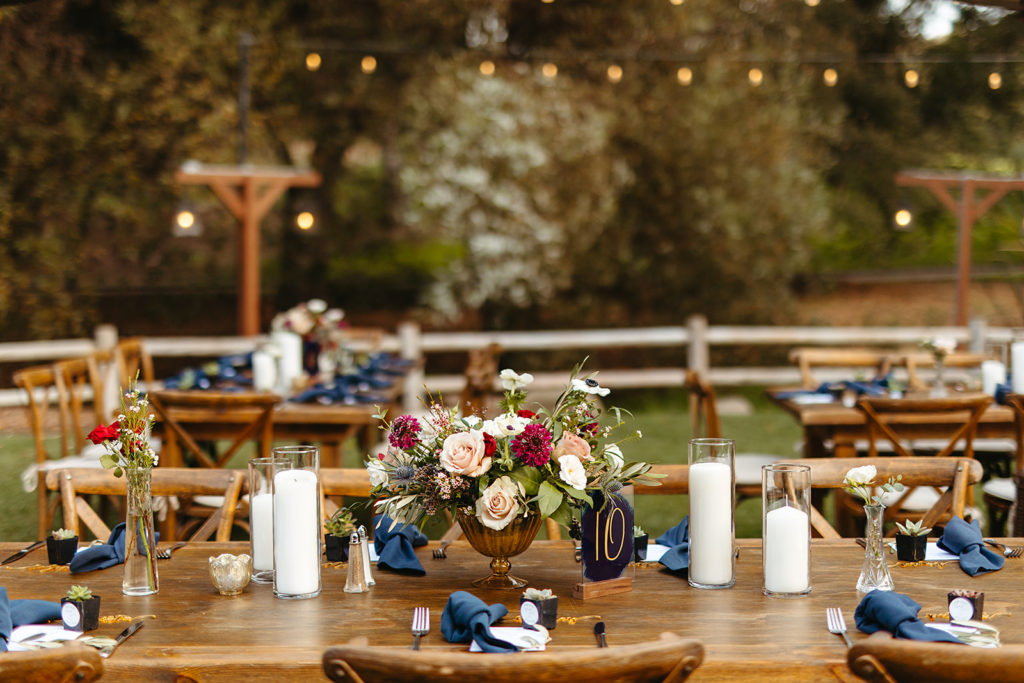 outdoor wedding reception at Temecula Creek Inn with wooden farmhouse tables, chairs and blue napkins with dark blue table numbers and floral arrangements with gold vases