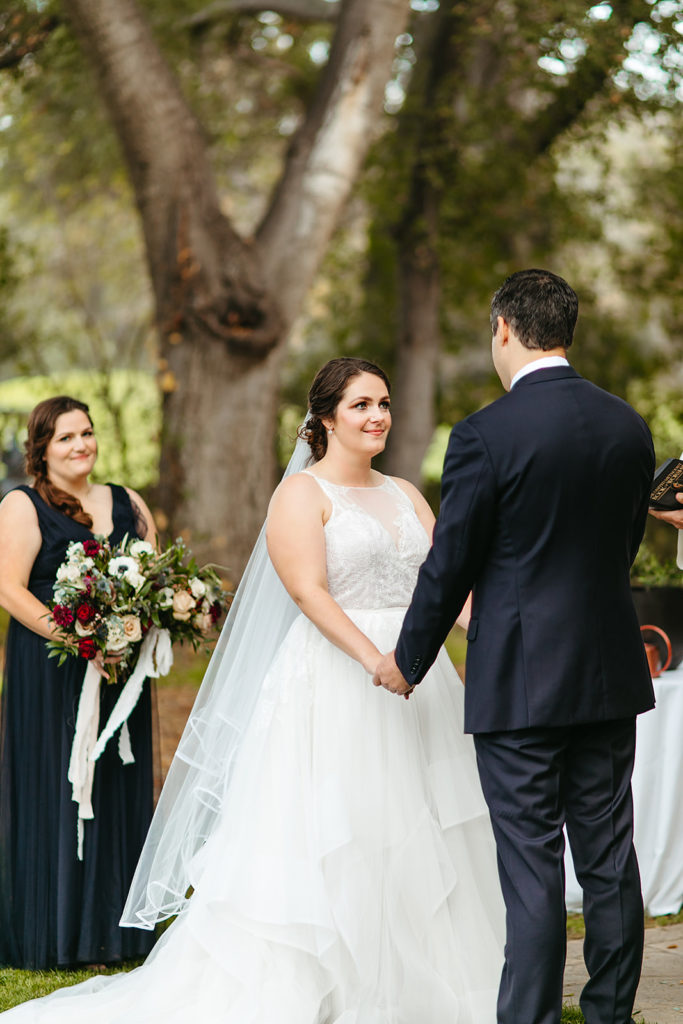 bride in tiered ballgown wedding dress holds hands with groom in navy suit and floral tie during hygge wedding ceremony at Temecula Creek Inn