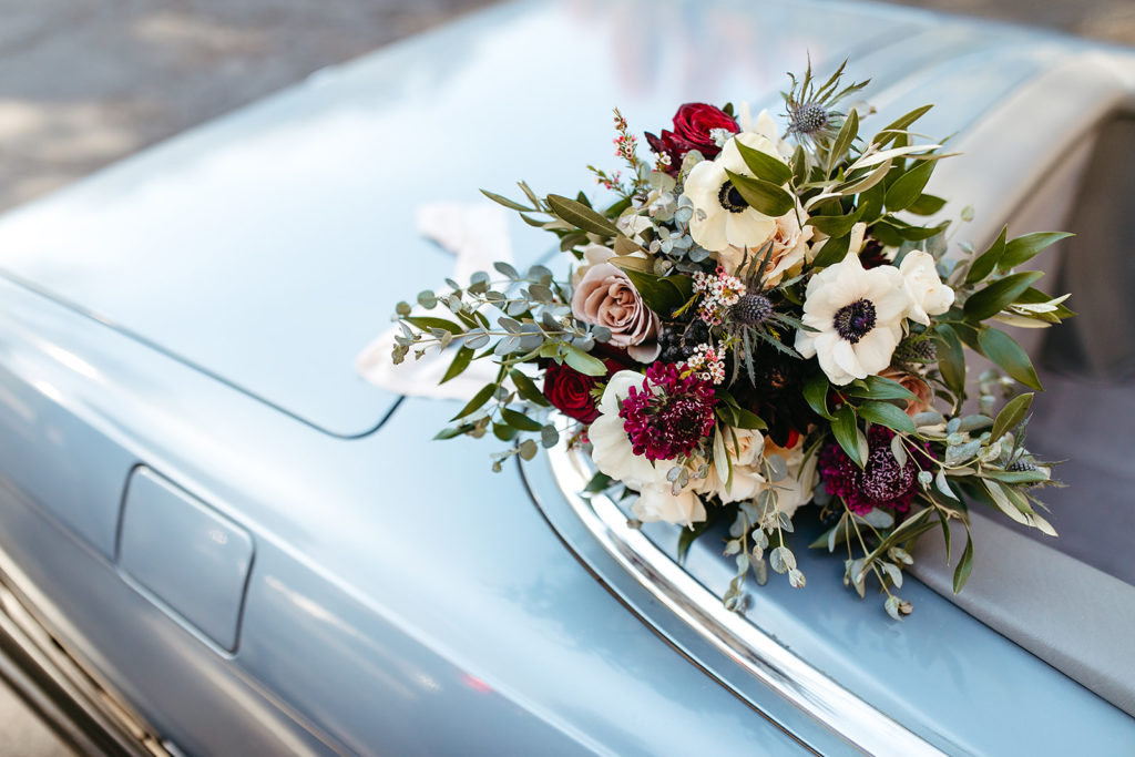 bridal bouquet with red roses, white anemone, blue sea holly and eucalyptus sits on top of vintage blue Mercedes 