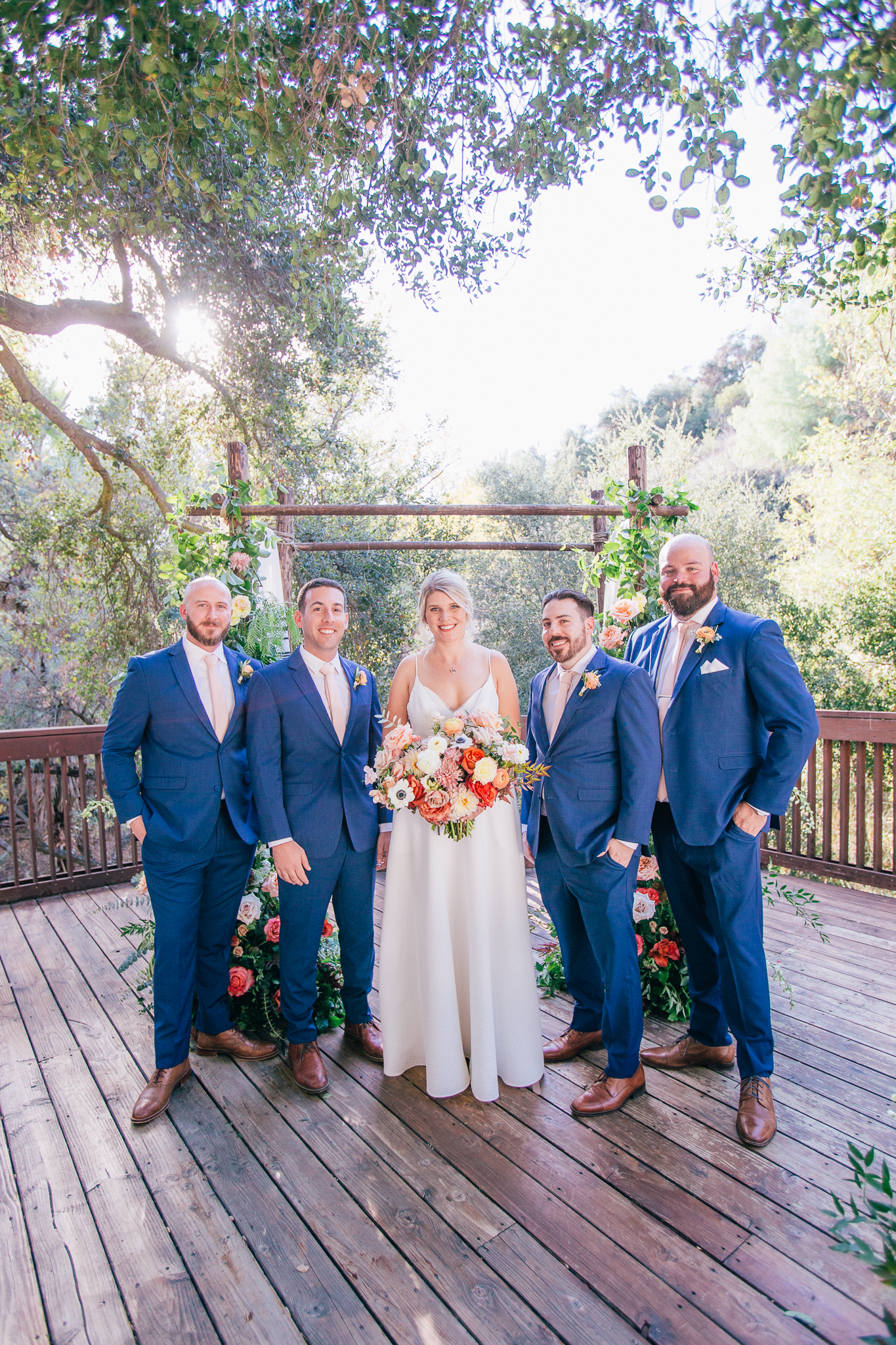 bride in spaghetti strap wedding dress stands with groomsmen in blue suits and light pink ties