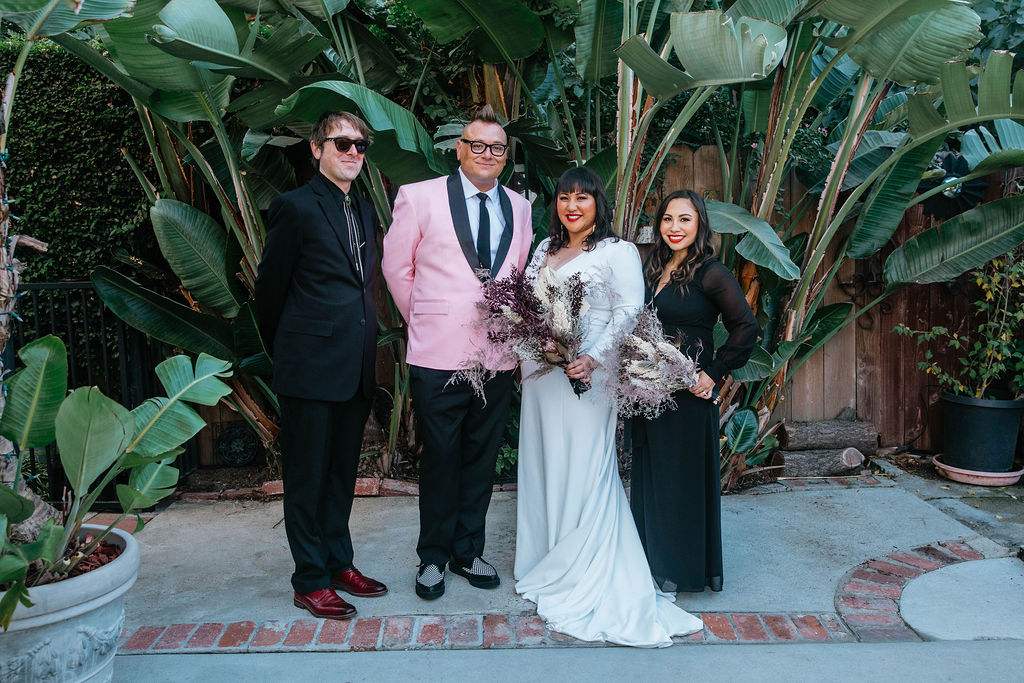 bride in long sleeve deep v-neck wedding dress holds bouquet of dried florals with groom in pink suit jacket and houndstooth shoes stand with with their wedding party in black suit and dress