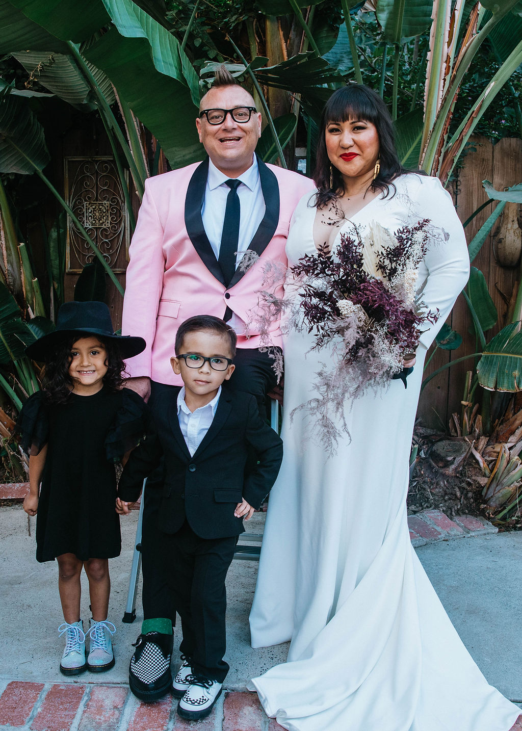 bride in long sleeve deep v-neck wedding dress holds bouquet of dried florals with groom in pink suit jacket and houndstooth shoes stand with with their flower child and ring bearer