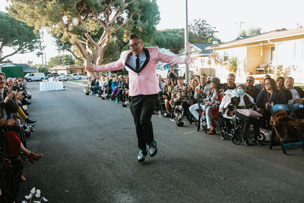 groom in pink suit jacket dances down wedding aisle under an urban canopy of trees in the middle of a residential street in Long Beach 