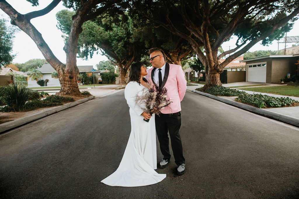 bride in long sleeve deep v-neck wedding dress holding bouquet of purple dried florals stands with groom in pink suit jacket and houndstooth shoes at ceremony site in the middle of a residential street under an urban canopy of trees