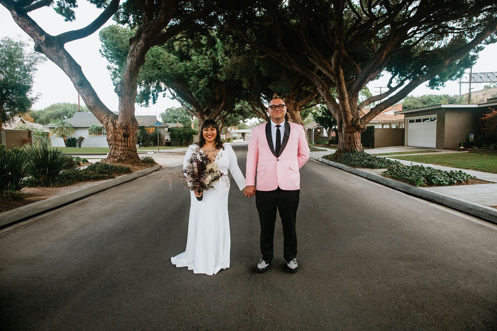 bride in long sleeve deep v-neck wedding dress holding bouquet of purple dried florals stands with groom in pink suit jacket and houndstooth shoes at ceremony site in the middle of a residential street under an urban canopy of trees