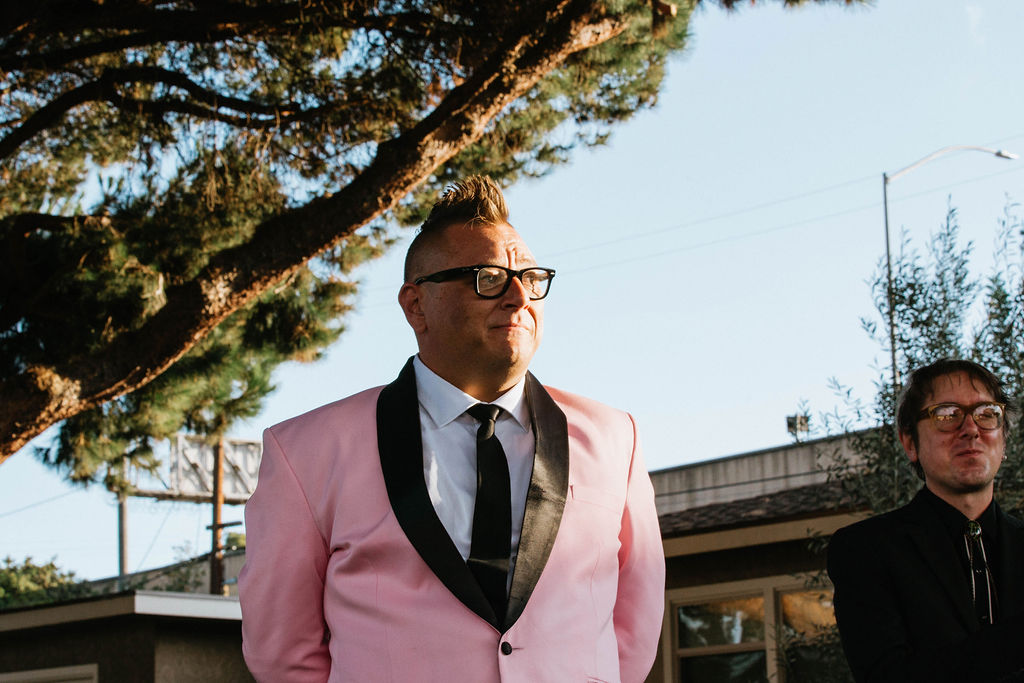 groom in pink suit jacket stands under urban canopy of trees during wedding ceremony