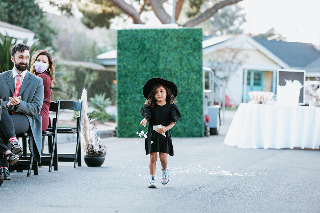 flower girl in black dress and hat walks down the aisle 