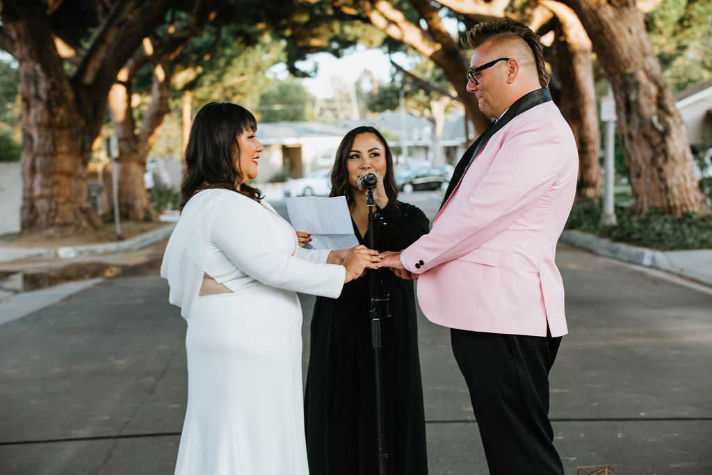 bride in long sleeve wedding dress holds hands with groom in pink suit jacket during wedding ceremony under an urban canopy of trees in the middle of a residential street in Long Beach