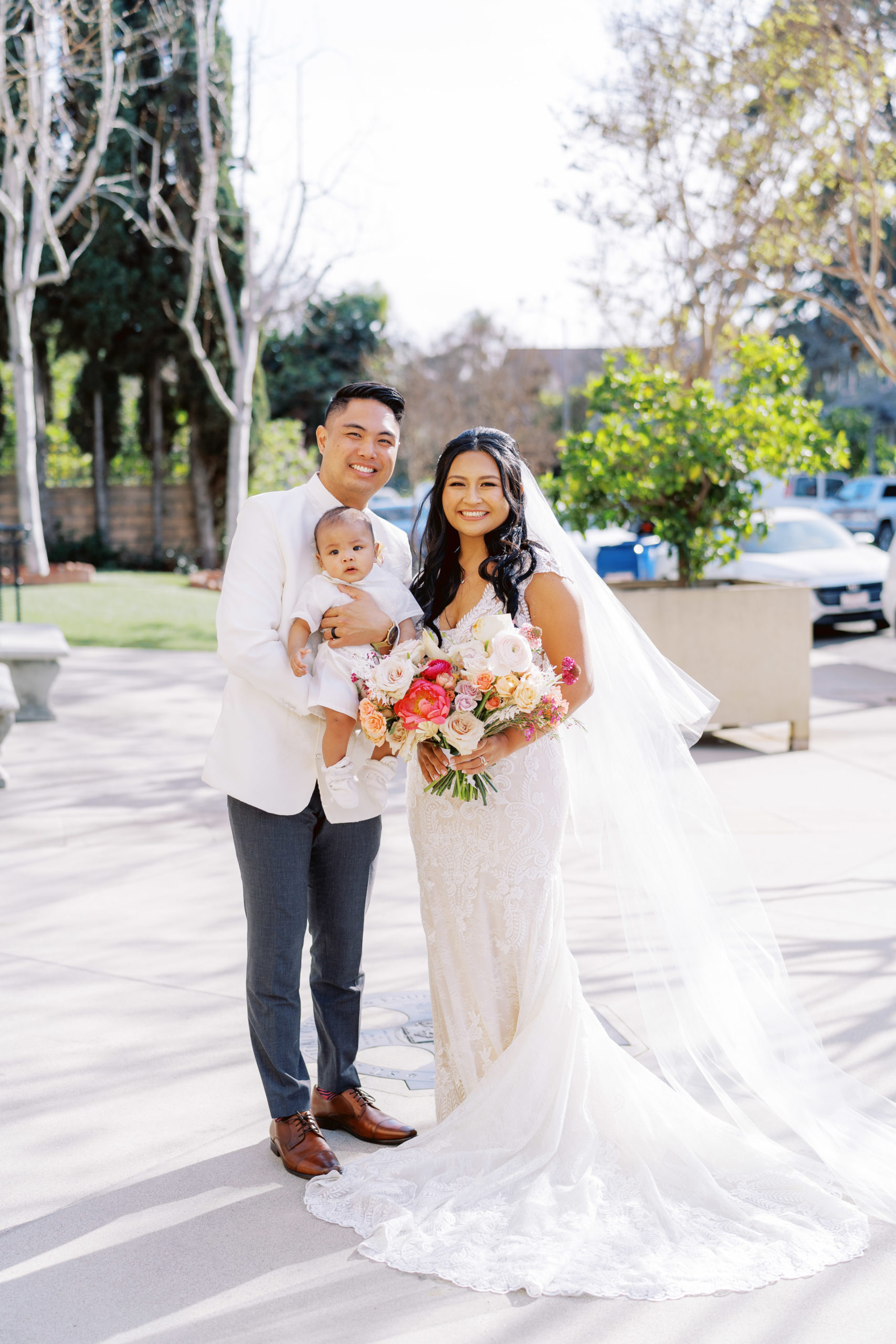 bride in lace wedding dress and bright bridal bouquet stands with groom in white wedding attire while holding baby during wedding portrait shots