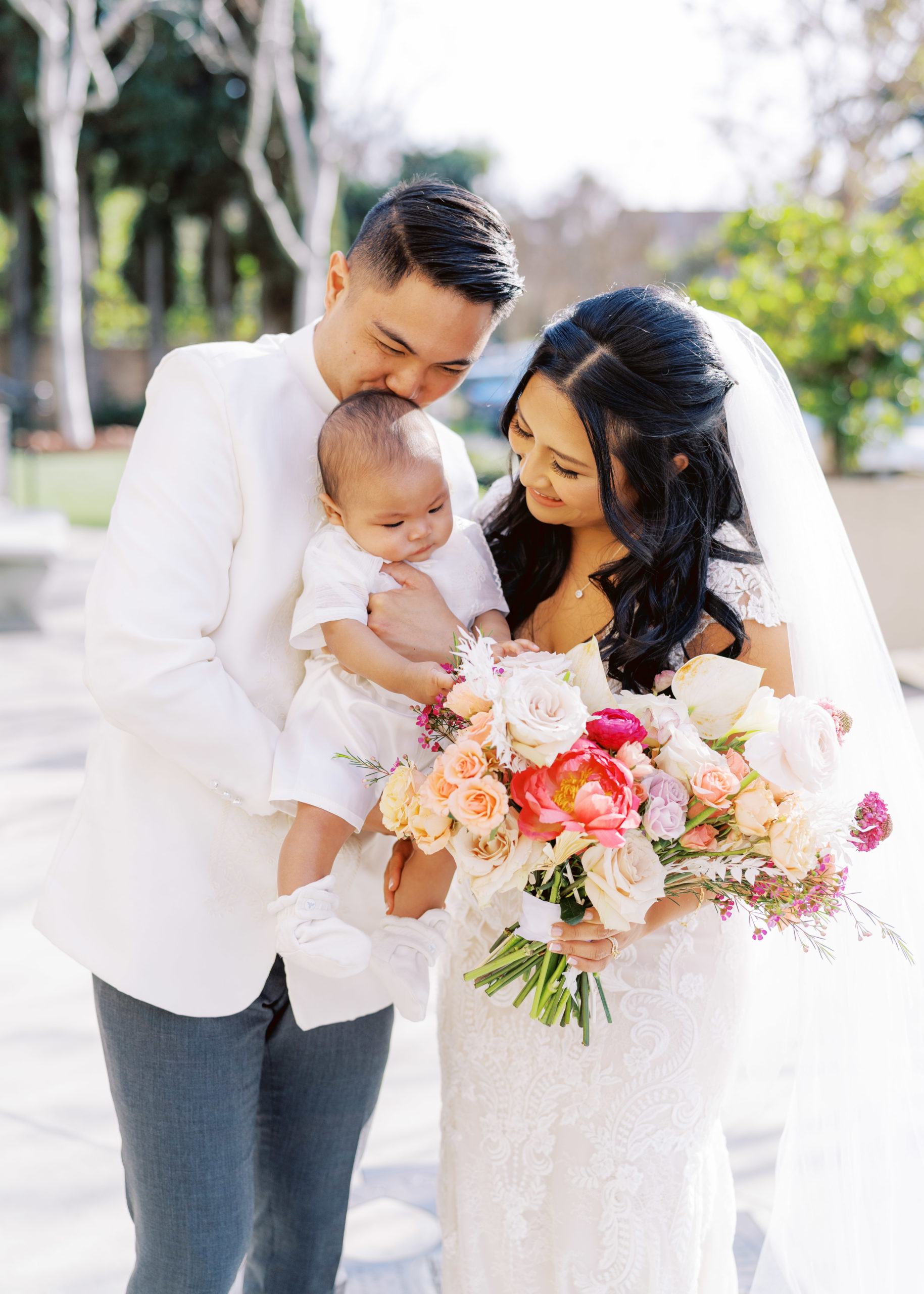 bride in lace wedding dress and bright bridal bouquet stands with groom in white wedding attire while holding baby during wedding portrait shots