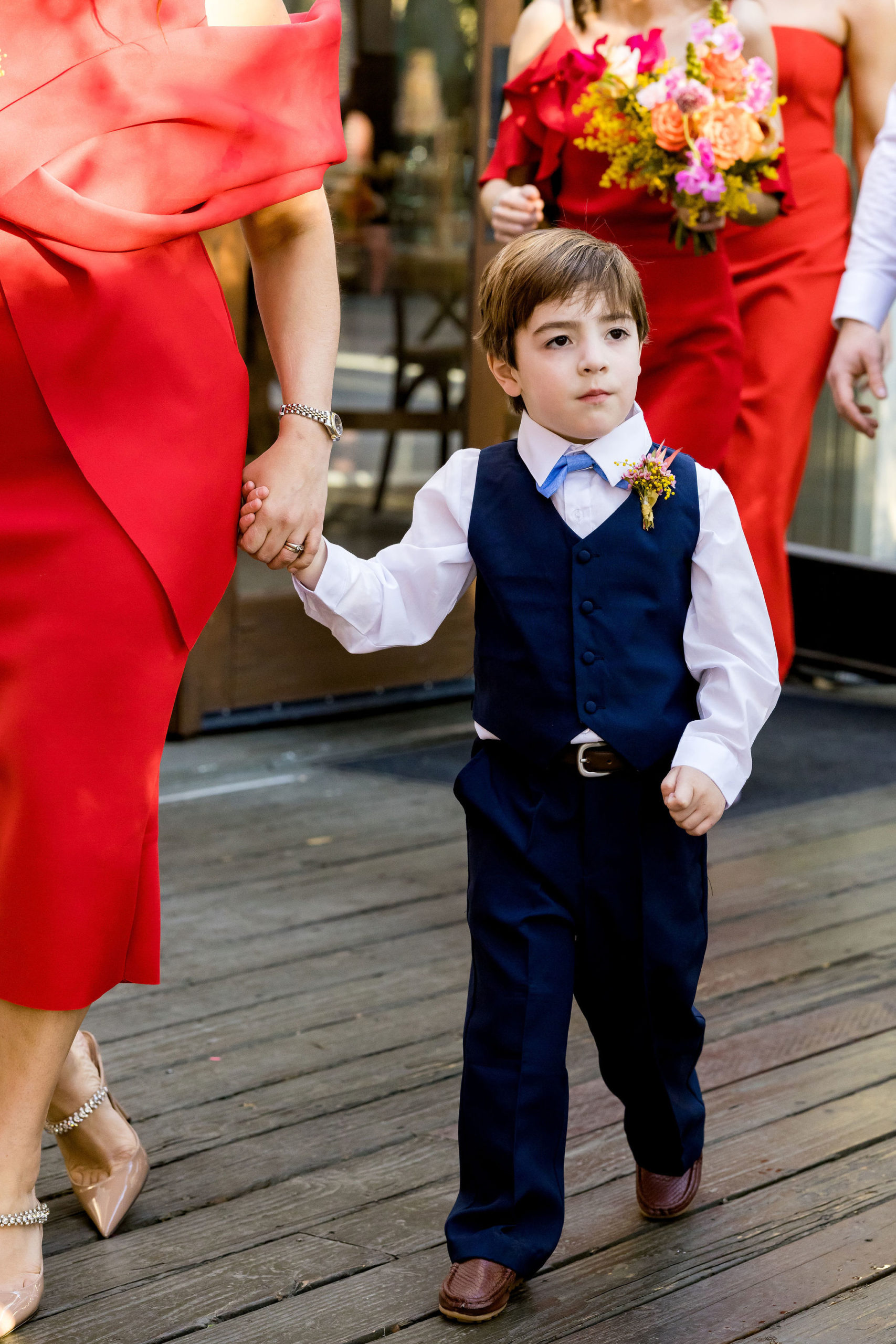 ring bearer with blue vest and bow tie walking down aisle with bridesmaids in red dresses