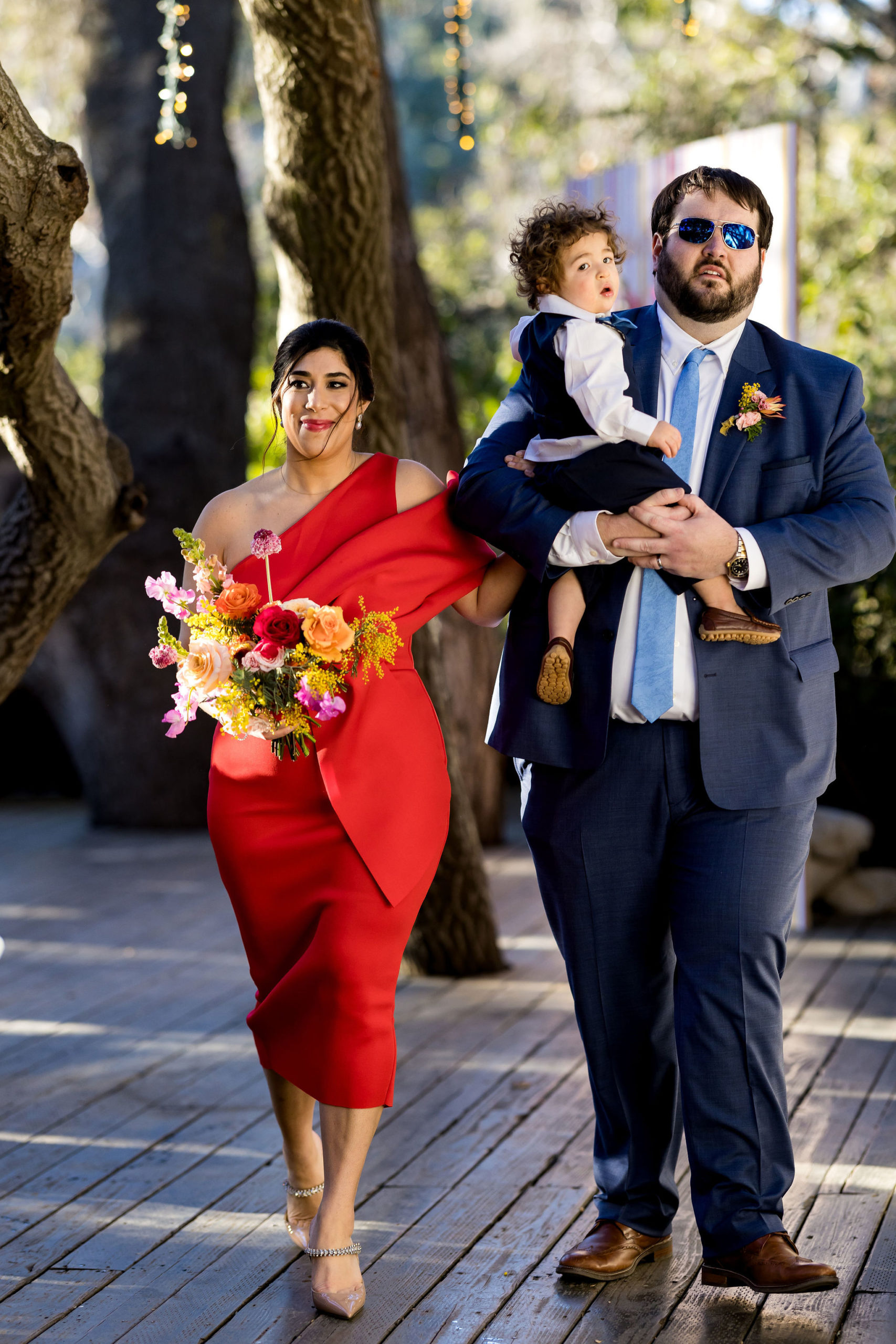 bridesmaid in modern red dress walks down aisle with groomsman in blue suit holding young ring bearer with blue vest