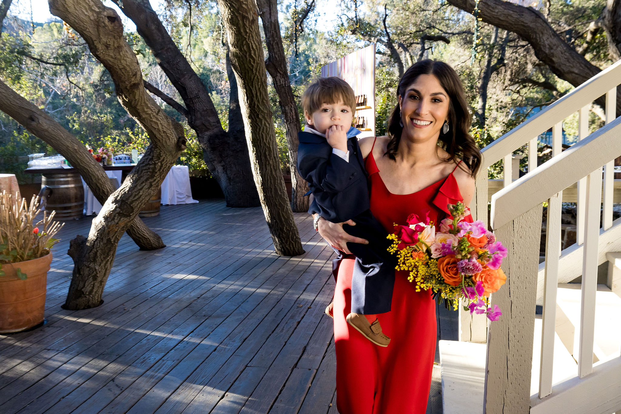 bridesmaid in red dress and bright floral bouquet holds young ring bearer with blue suit and bowtie 