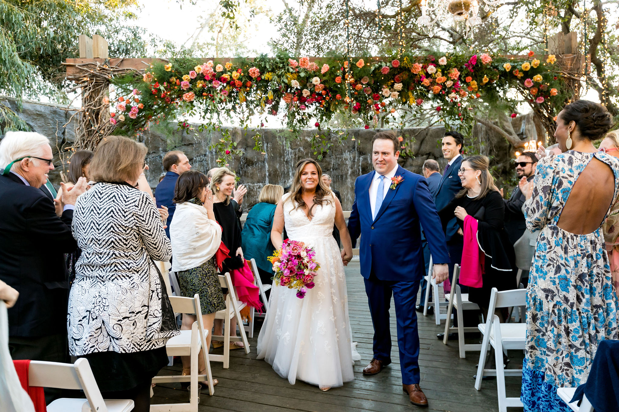 bride and groom recessional during bright and energetic wedding ceremony at Calamigos Ranch with red, pink, orange, yellow, purple and white florals hanging in front of waterfall 