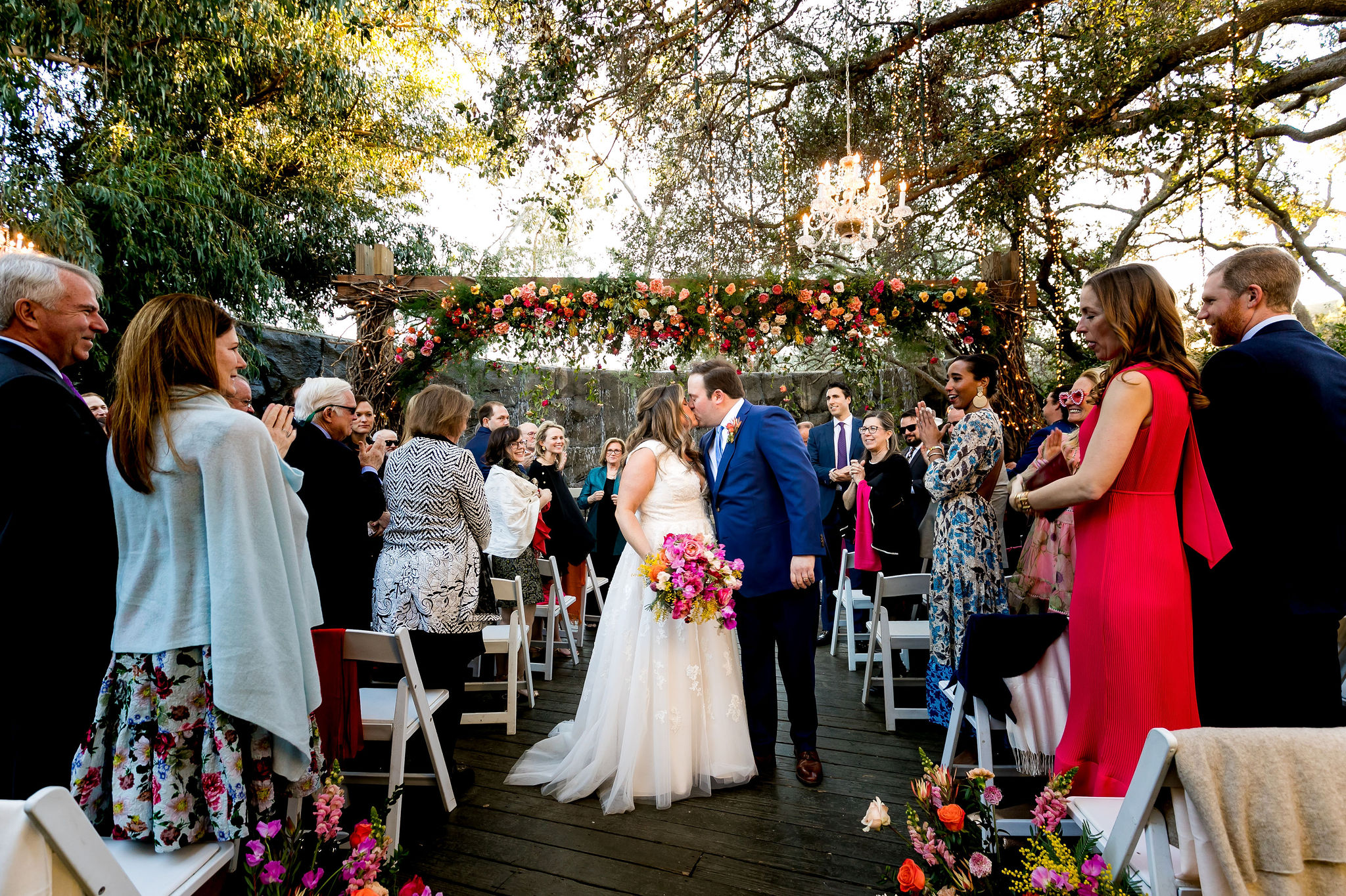 bride and groom kiss during bright and energetic wedding ceremony at Calamigos Ranch with red, pink, orange, yellow, purple and white florals hanging in front of waterfall 
