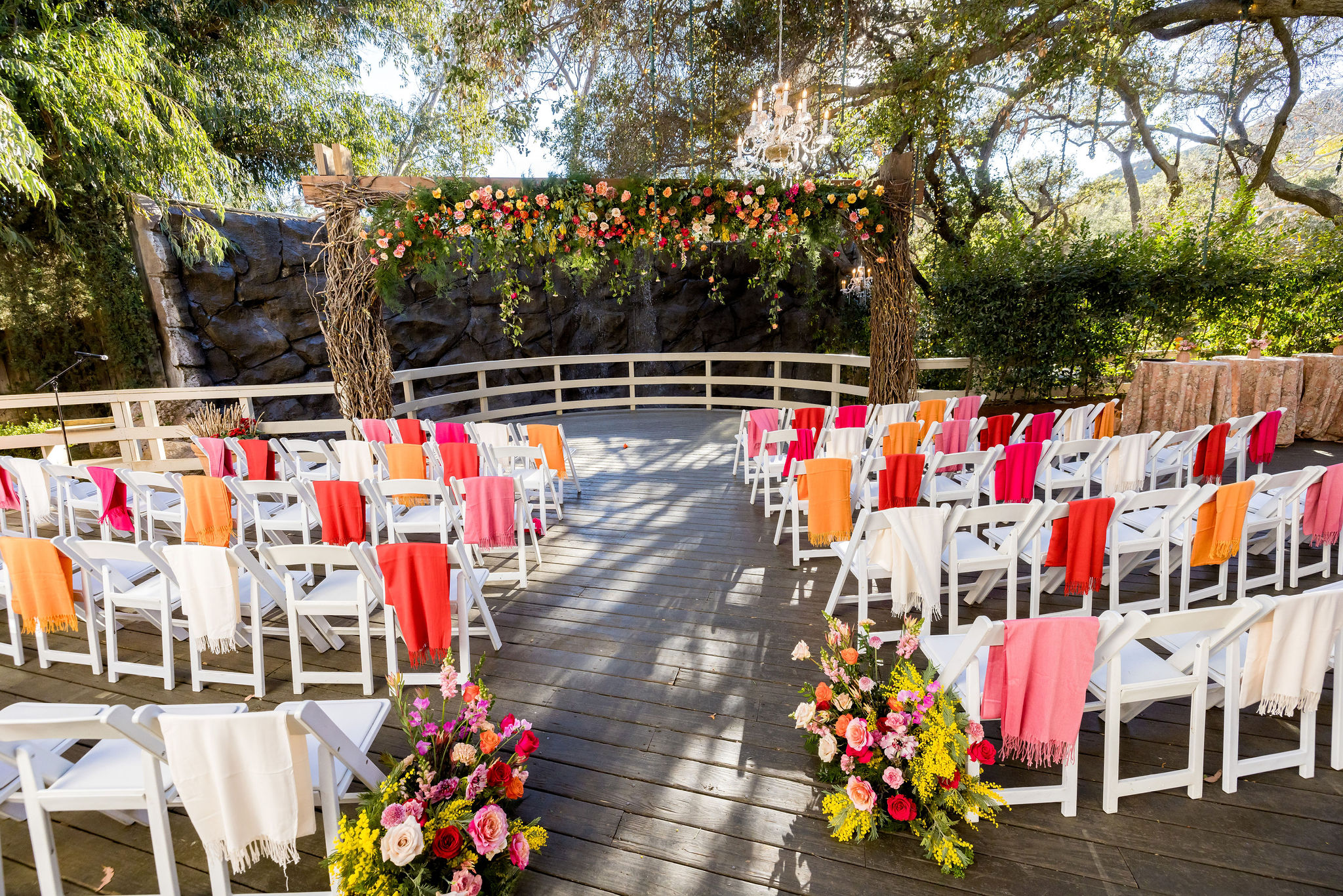 An energetic wedding ceremony at the Oak Room at Calamigos Ranch ceremony space with bright orange, red, pink and white florals and shawls hanging from ceremony chairs