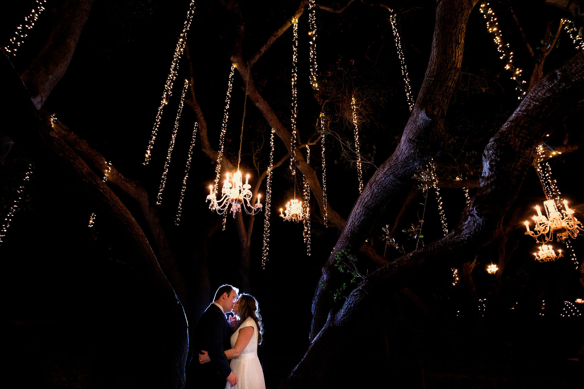 night time photo of bride and groom kissing under chandeliers and sparkling lights in trees