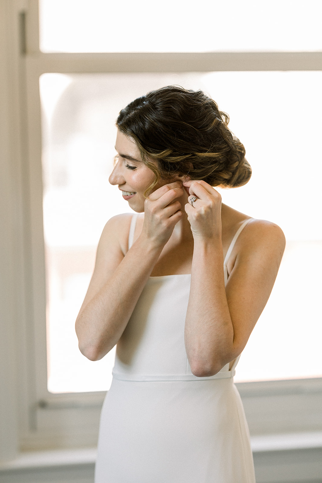 bride in contemporary spaghetti strap dress and low chignon bun puts on jewelry 