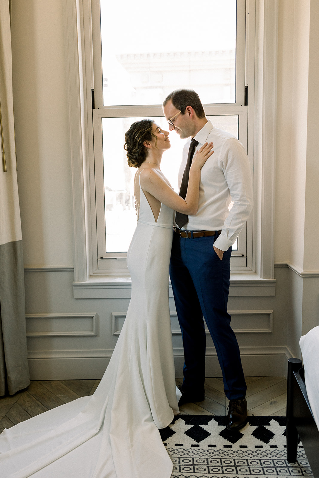 bride in contemporary spaghetti strap dress and low chignon bun poses with groom wearing glasses and white shirt with navy pants