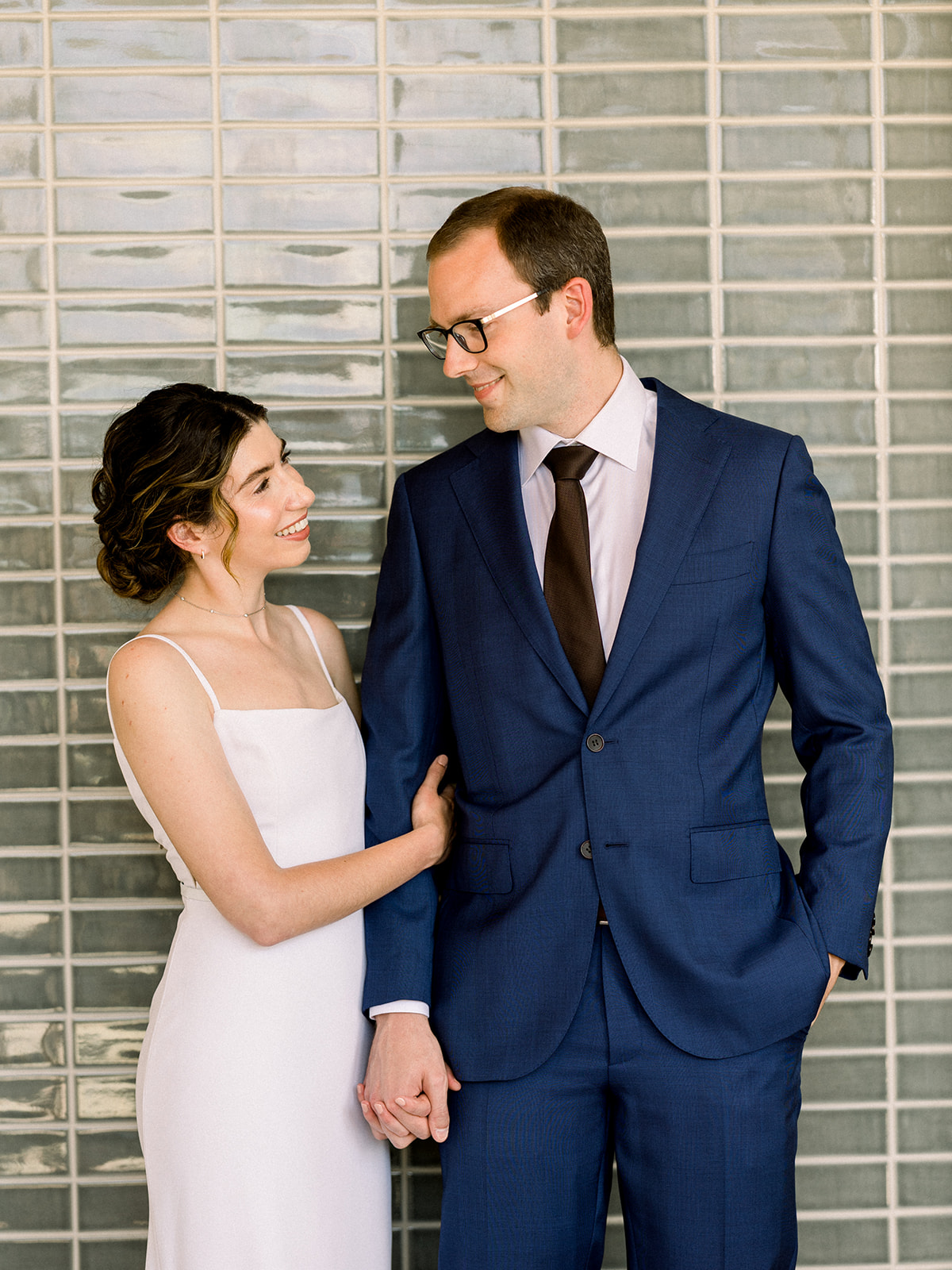 bride in contemporary spaghetti strap dress and low chignon bun poses with groom wearing glasses and navy suit