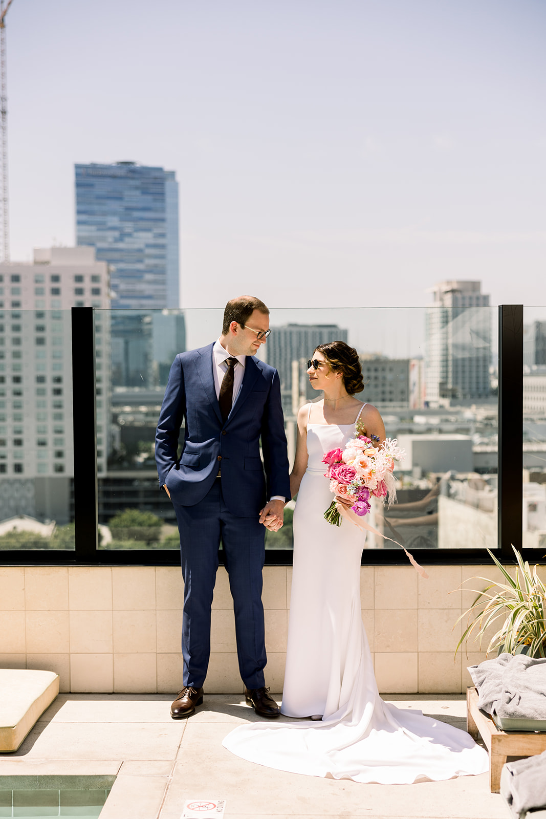 bride in contemporary spaghetti strap dress and groom in navy suit with black tie stand on rooftop with DTLA skyline in background