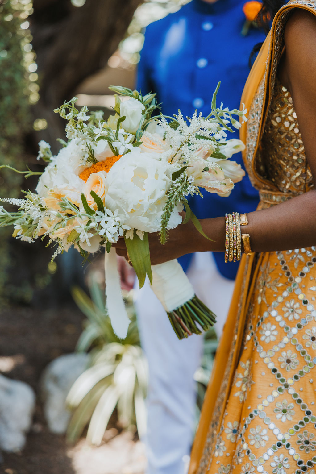 bride in orange marigold colored wedding sari holds bouquet of white and orange flowers during first look with groom in cobalt blue suit and orange marigold boutonniere at Lake Shrine Temple
