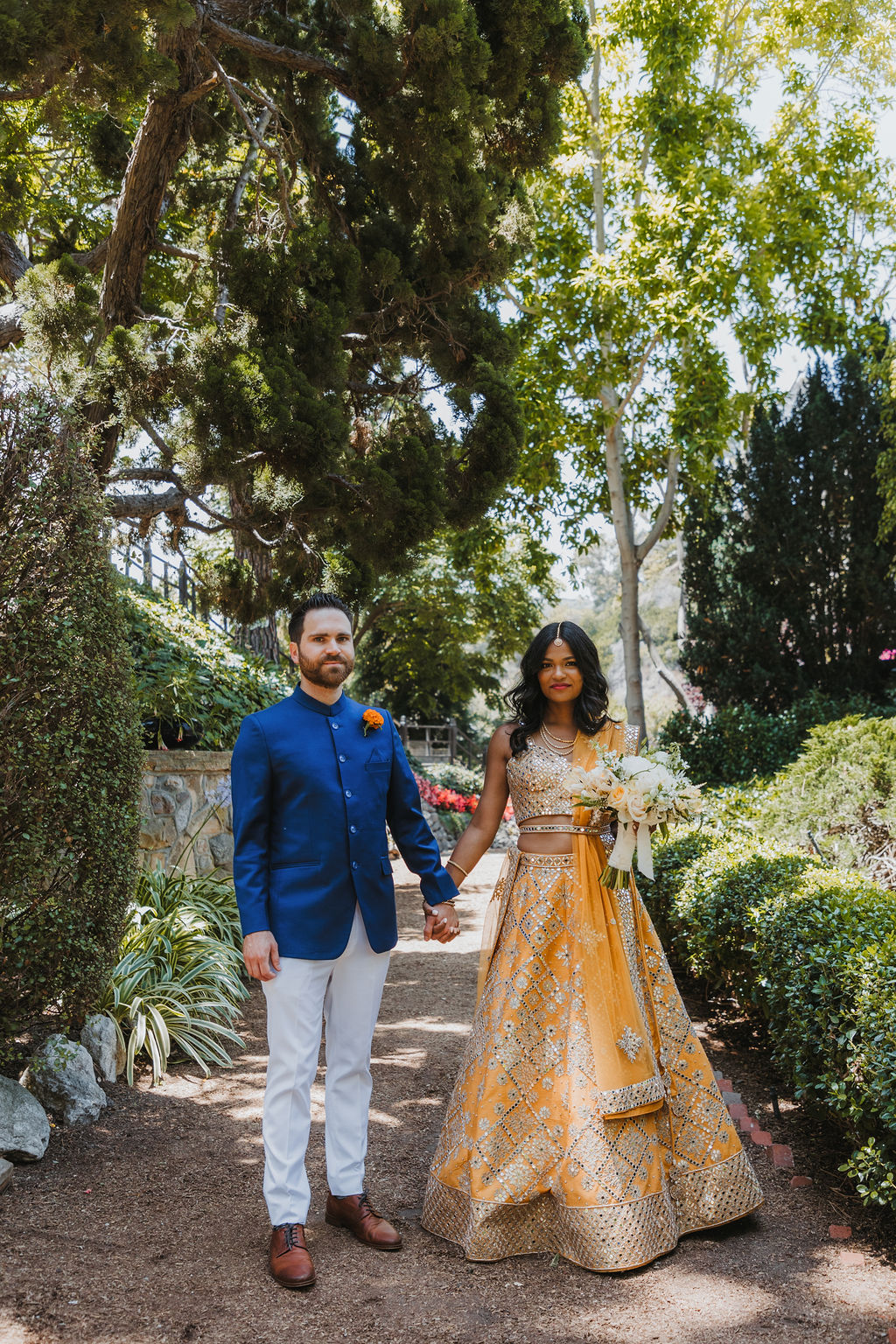 bride in orange marigold colored wedding sari holds bouquet of white and orange flowers during portraits with groom in cobalt blue suit and orange marigold boutonniere at Lake Shrine Temple