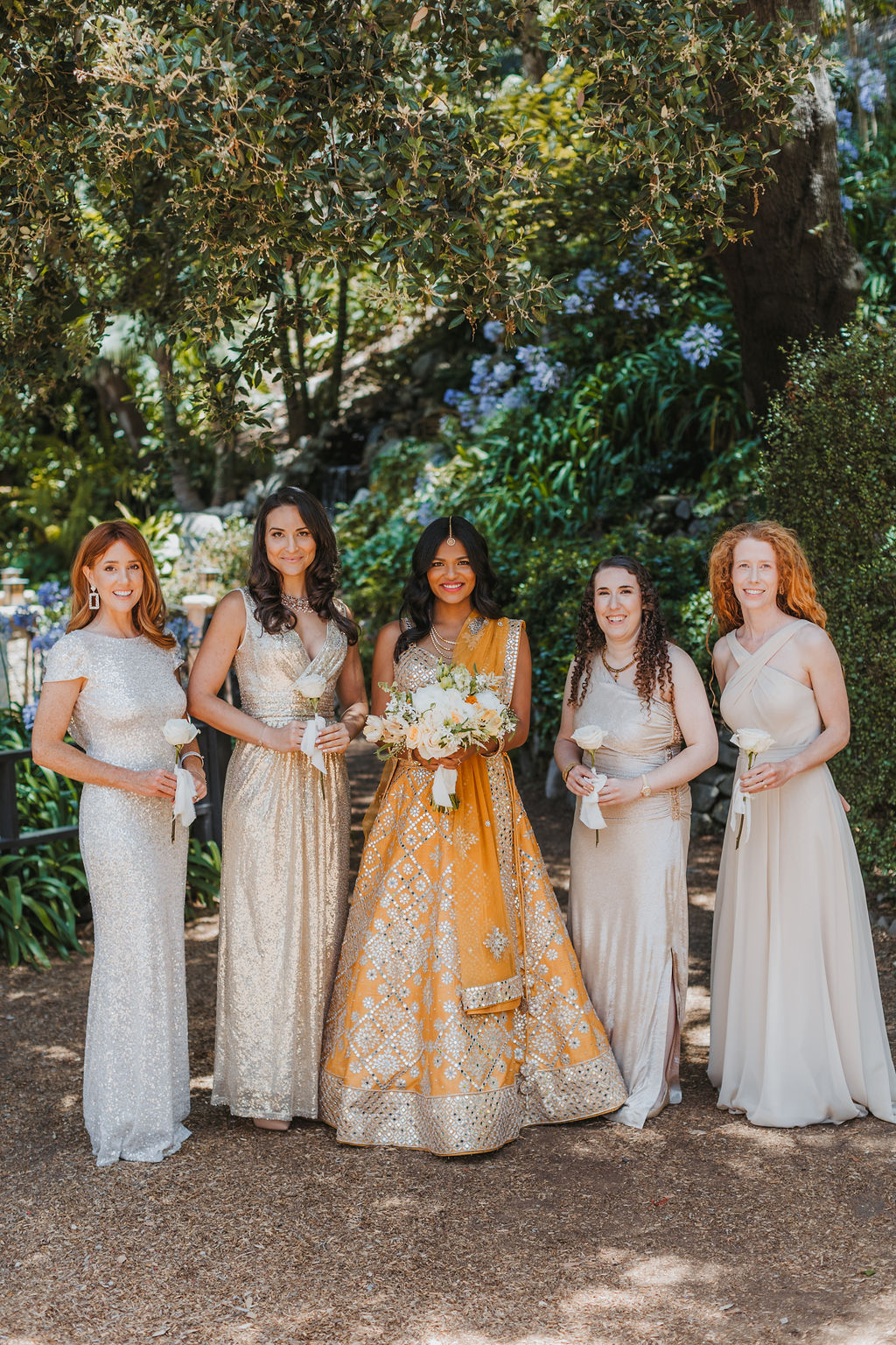 bride in orange marigold colored wedding sari holds bouquet of white and orange flowers during portraits with bridesmaids in mix matched gold and beige bridesmaid dresses