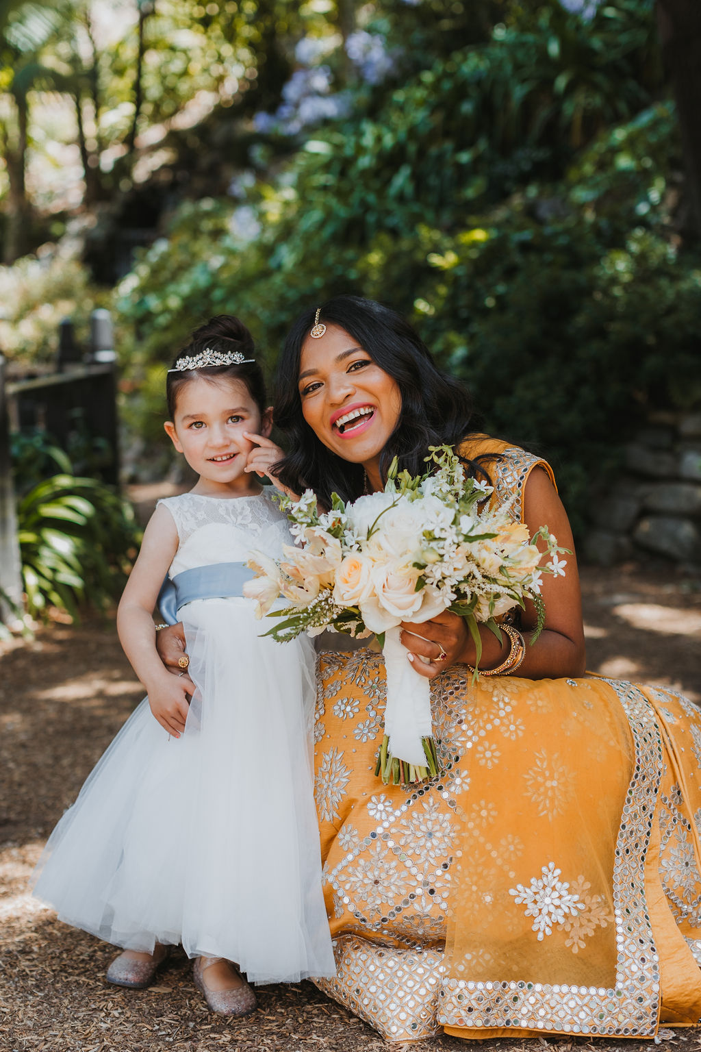 bride in orange marigold colored wedding sari holds bouquet of white and orange flowers during portraits with flower girl in white dress and tiara