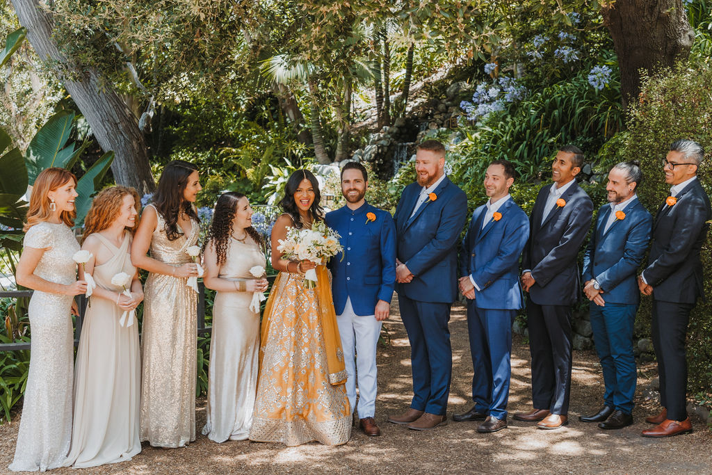 bride in orange marigold colored wedding sari holds bouquet of white and orange flowers with groom in cobalt blue suit stands with wedding party wearing mix matched gold and beige bridesmaid dresses and blue and grey suits at Lake Shrine Temple