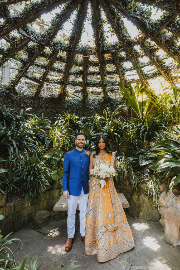 bride wearing orange sari with white and orange bouquet stands with groom wearing cobalt blue suit jacket and orange marigold boutonniere underneath an open air greenhouse at Lake Shrine