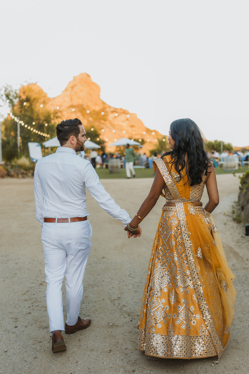 bride in orange sari walks with groom in white to wedding reception at Saddlerock Ranch in Malibu