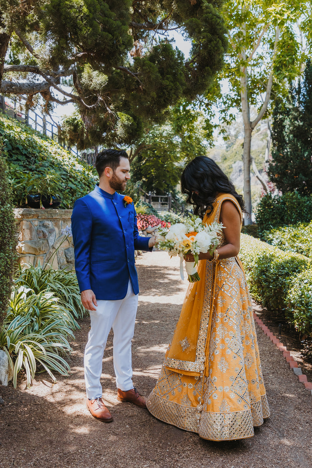 bride in orange marigold colored wedding sari holds bouquet of white and orange flowers during first look with groom in cobalt blue suit and orange marigold boutonniere at Lake Shrine Temple