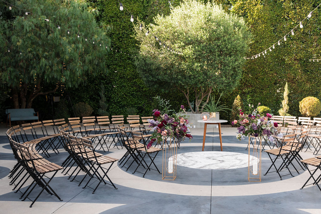 circle outdoor wedding ceremony seating at The Fig House courtyard with two gold geometric candle holders with pink and purple floral arrangements on tops