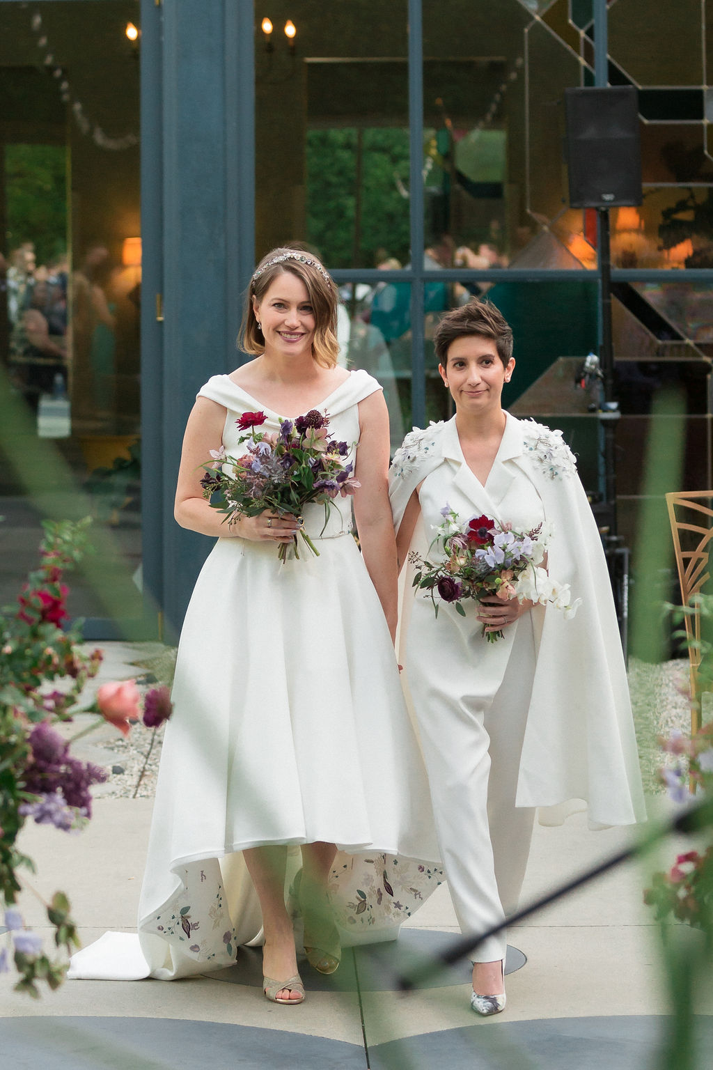two brides hold hands walking down wedding aisle in custom made coordinated wedding attire with one bride wearing a hi low two piece wedding dress and the other bride in white bridal jumpsuit and embellished cape 