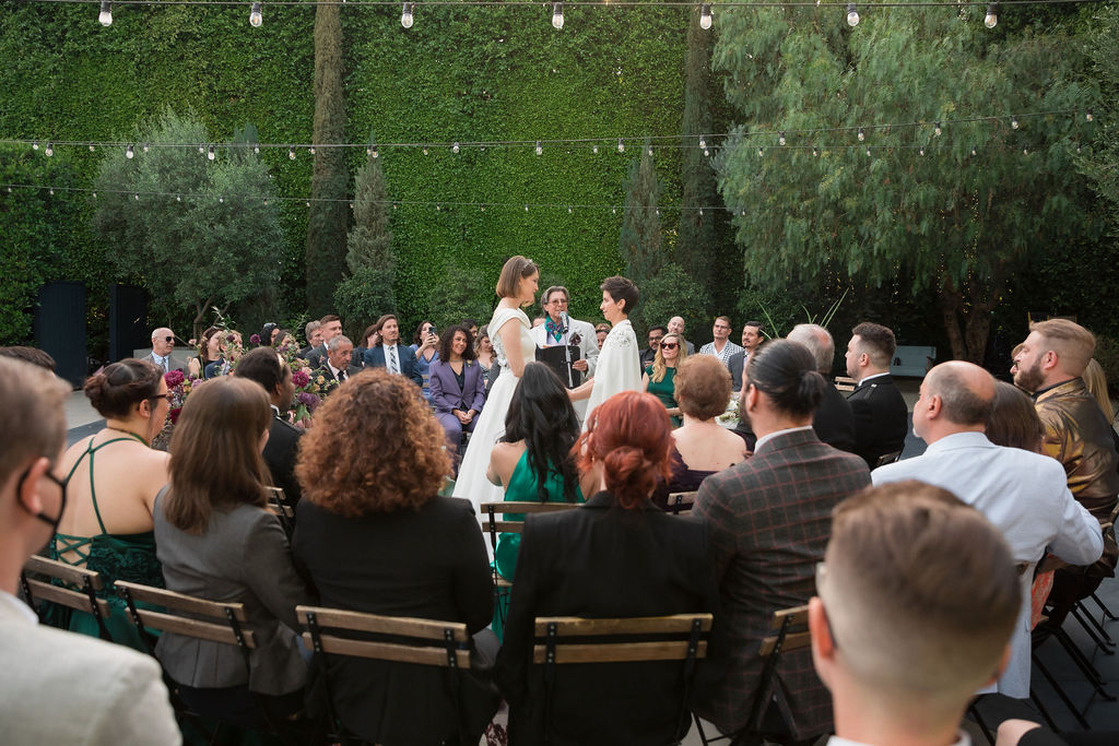 two brides wearing coordinating bridal attire hold hands during outdoor wedding ceremony at the Fig House