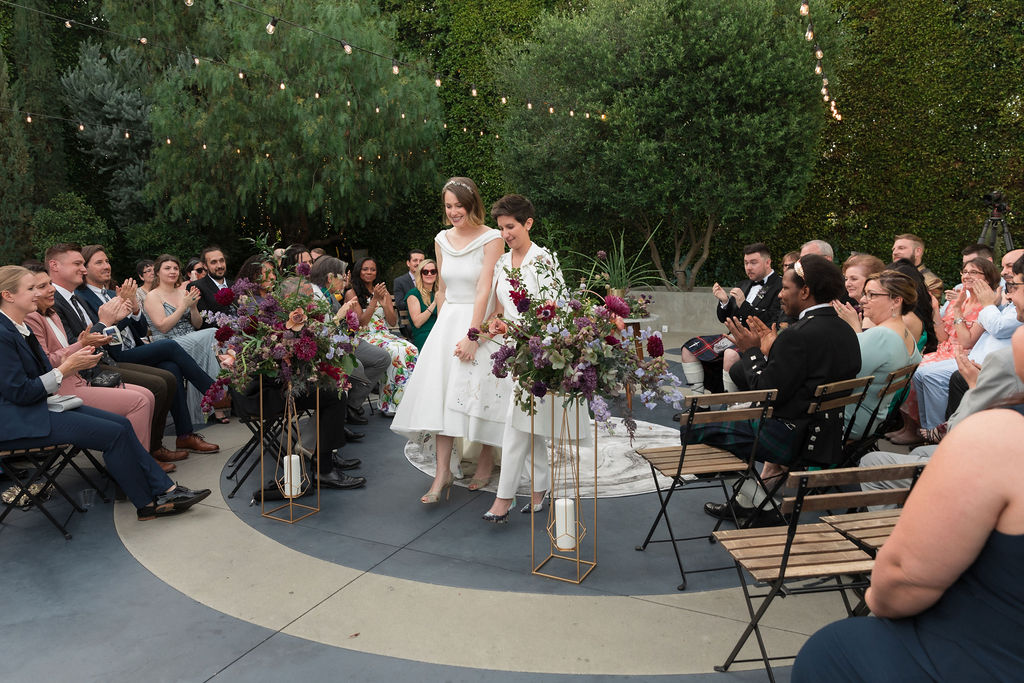 two brides wearing coordinating bridal attire hold hands during recessional of outdoor wedding ceremony at the Fig House