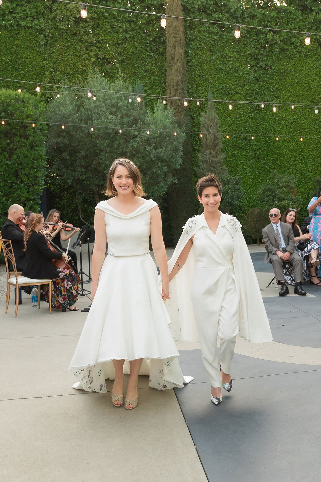 two brides wearing coordinating bridal attire hold hands during recessional of outdoor wedding ceremony at the Fig House