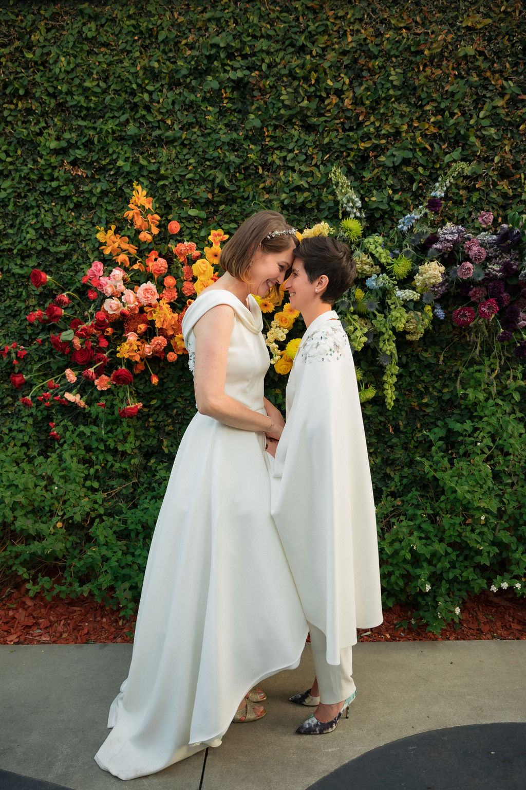 brides in coordinating bridal dress and bridal jumpsuit hold hands in front of hanging rainbow floral installation after wedding ceremony at The Fig House